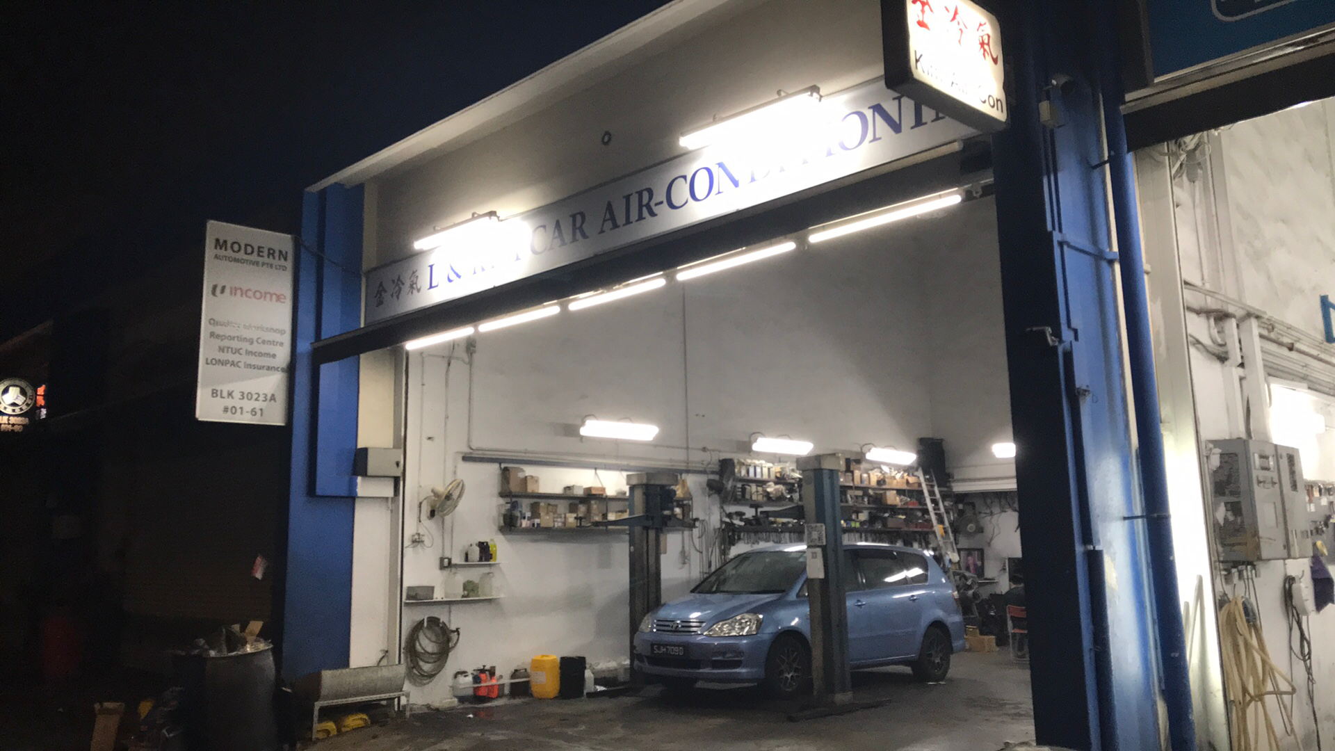 The L and Kim Car Air-Conditioning workshop lit up at night on Ubi Road 1, with a car raised on the two-post lift inside