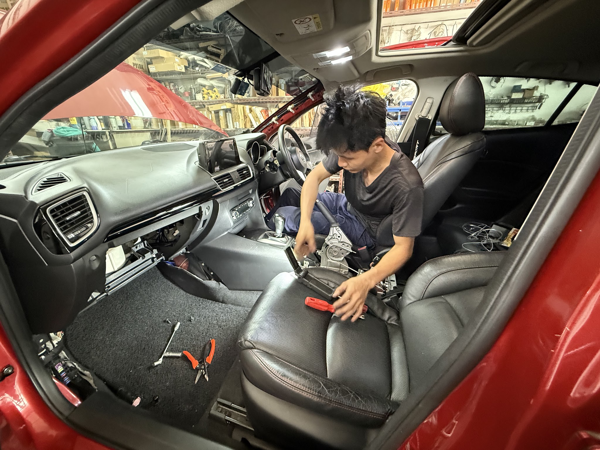 A technician working inside a red car with the front passenger seat removed to reach the aircon system